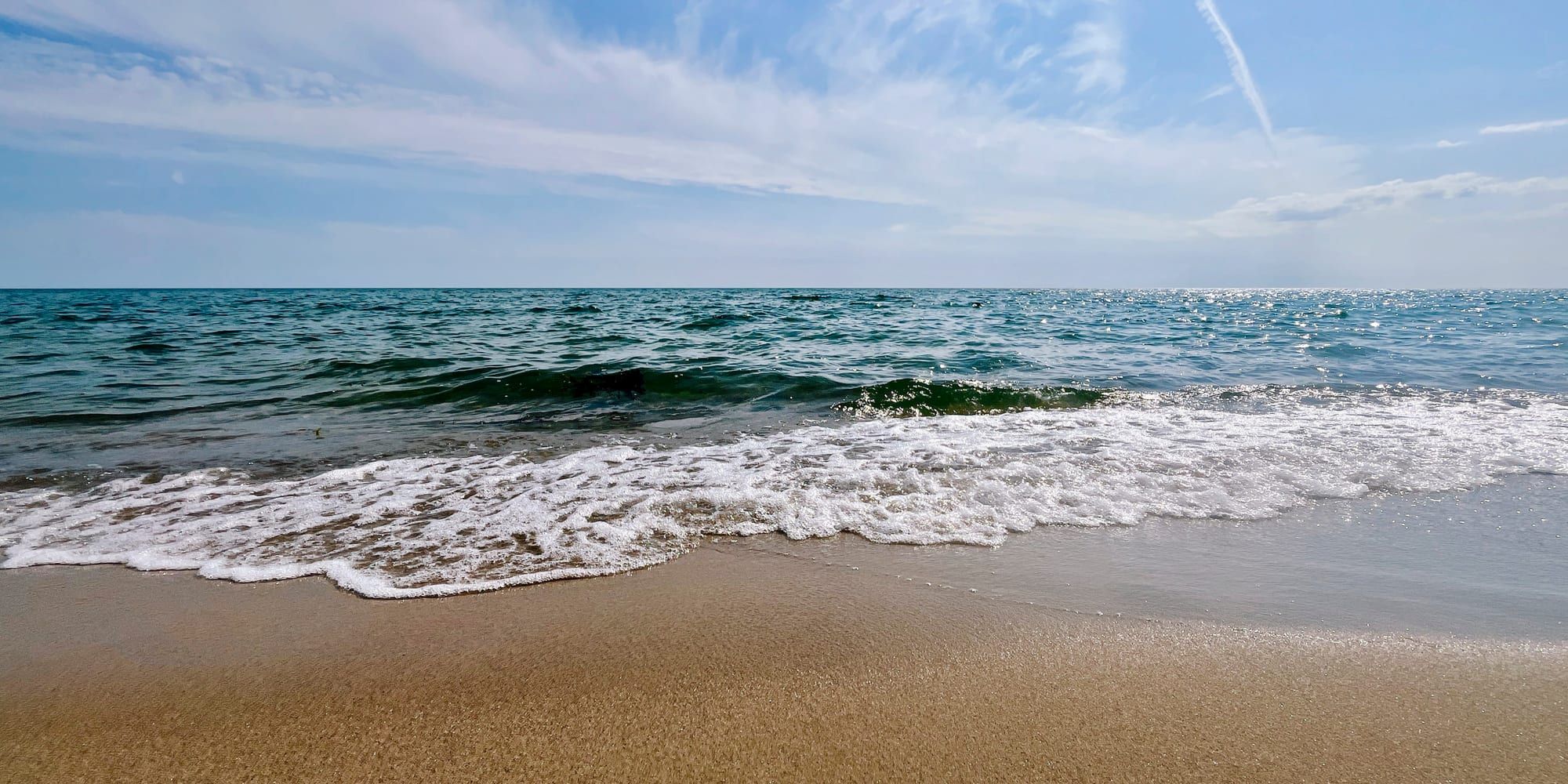 Small waves breaking onto a wide sandy beach on a bright summer day, foam spreading thin across the sand in the foreground, the sea flat and glittering out to a hazy horizon under a pale blue sky with thin high cloud.