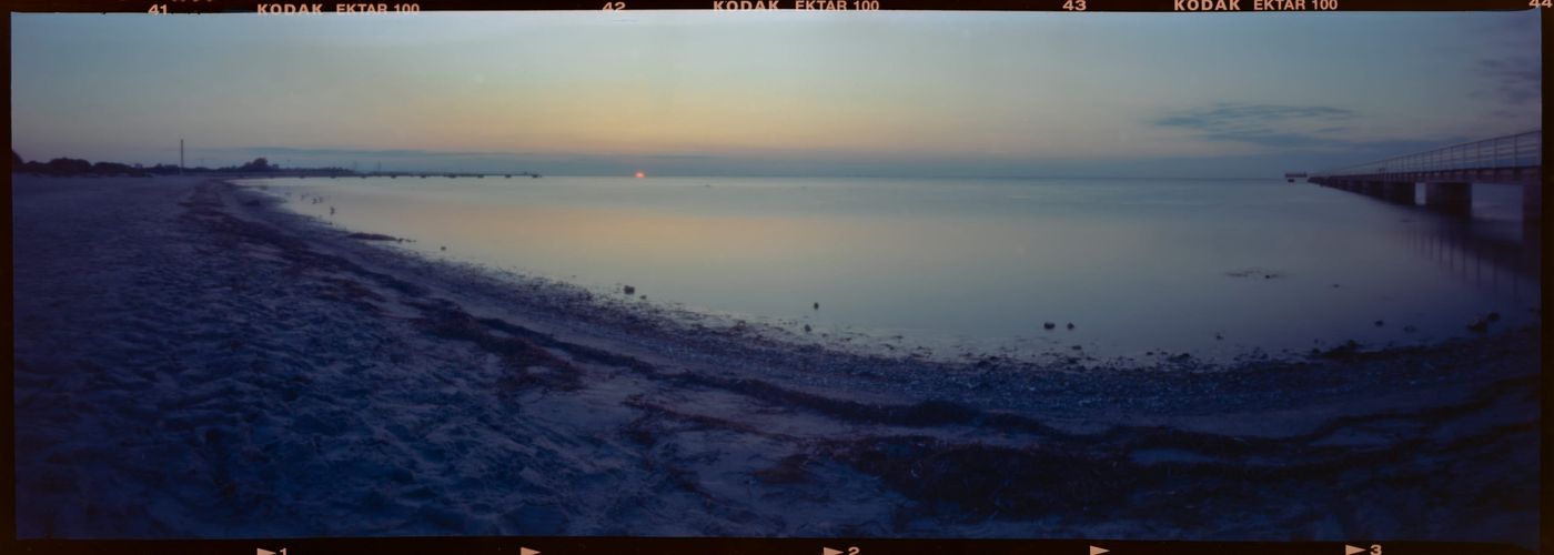 A panoramic film frame at dusk — a calm, glassy sea under a soft pastel sky, the sun a small point low on the horizon at centre, a sandy beach in the foreground crossed by a curving band of dark seaweed, and a long low pier with railings reaching out into the water on the right. The black film rebate with “Kodak Ektar 100” edge markings frames the image.