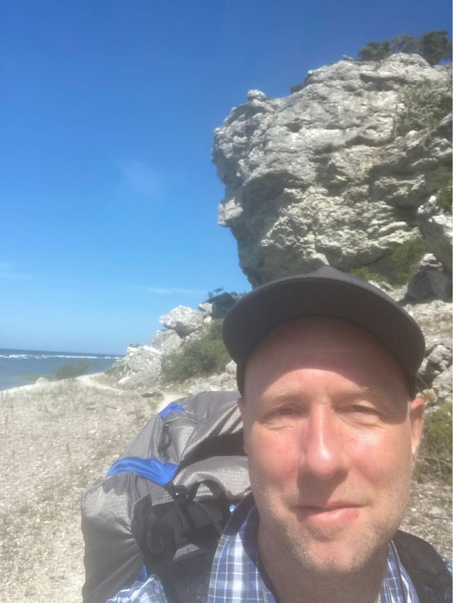 Selfie av en vandrare i keps och rutig skjorta med en stor vandringsryggsäck, en kalkstensklippa och havet bakom.